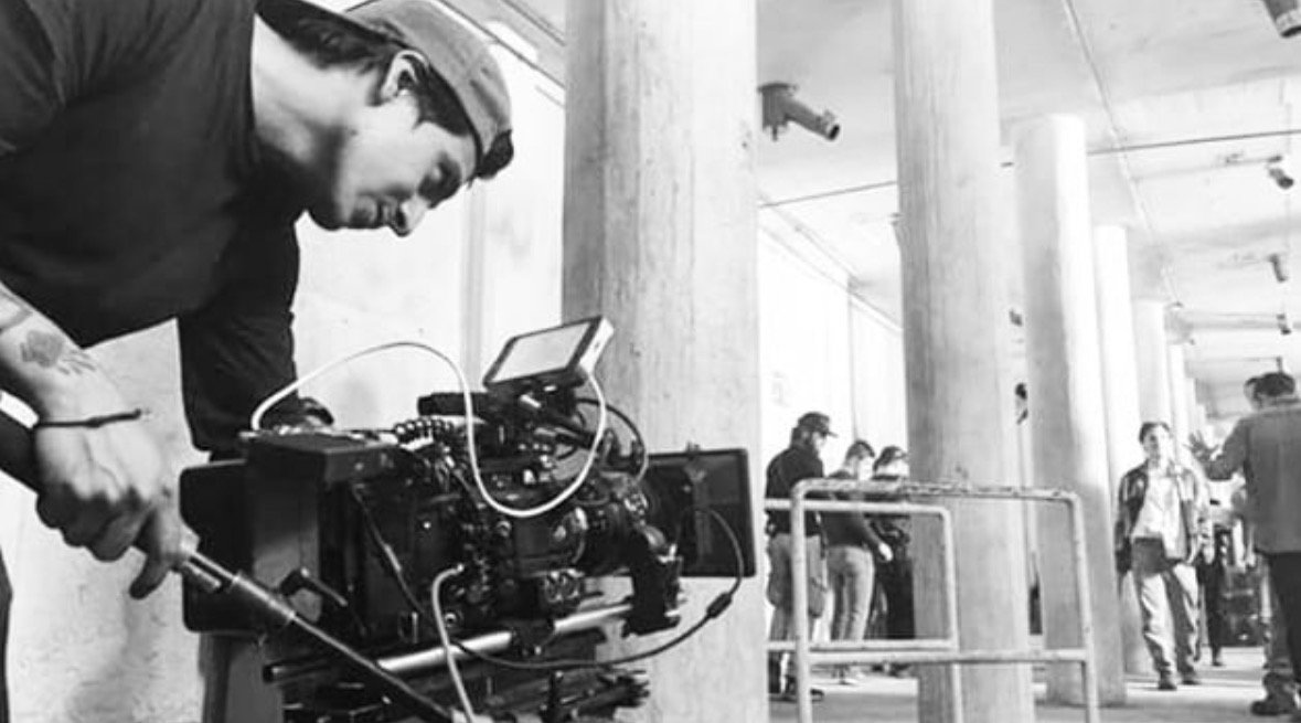 Juan Fernando operando una cámara de cine en un set con columnas, con el equipo de producción al fondo